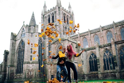 young couple smiling and throwing Autumn leaves into the air with Hereford Cathedral behind them